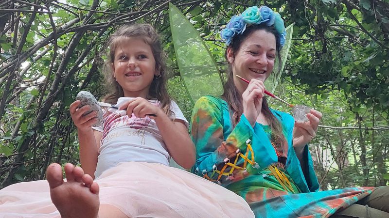 Eine Frau und ein Mädchen als Feen im Wald sitzend, mit Steinen und Zauberstäben.